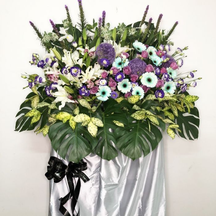 Elegant condolence wreath featuring white lilies, purple roses, and blue gerberas arranged with tropical foliage and a black ribbon, symbolizing sympathy and respect.