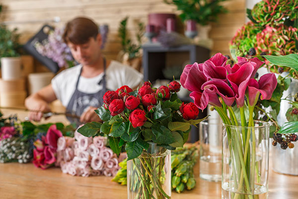 Florist working in a shop with fresh and preserved flowers, including red roses and pink calla lilies, arranged in glass vases on a wooden counter.
