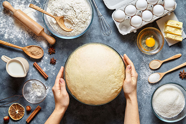 Top-down view of baking ingredients and tools surrounding a bowl of risen dough held by two hands, including flour, eggs, butter, sugar, milk, cinnamon sticks, star anise, and utensils on a dark countertop.