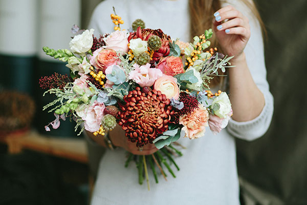 Person holding a rustic, hand-tied bouquet made of various fresh flowers including roses, chrysanthemums, eucalyptus, and berries in soft and warm tones.