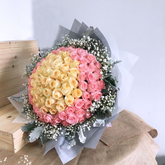 Large rose bouquet Singapore featuring a stunning blend of yellow and pink roses, accented with baby’s breath and silver foliage, wrapped in soft white paper.