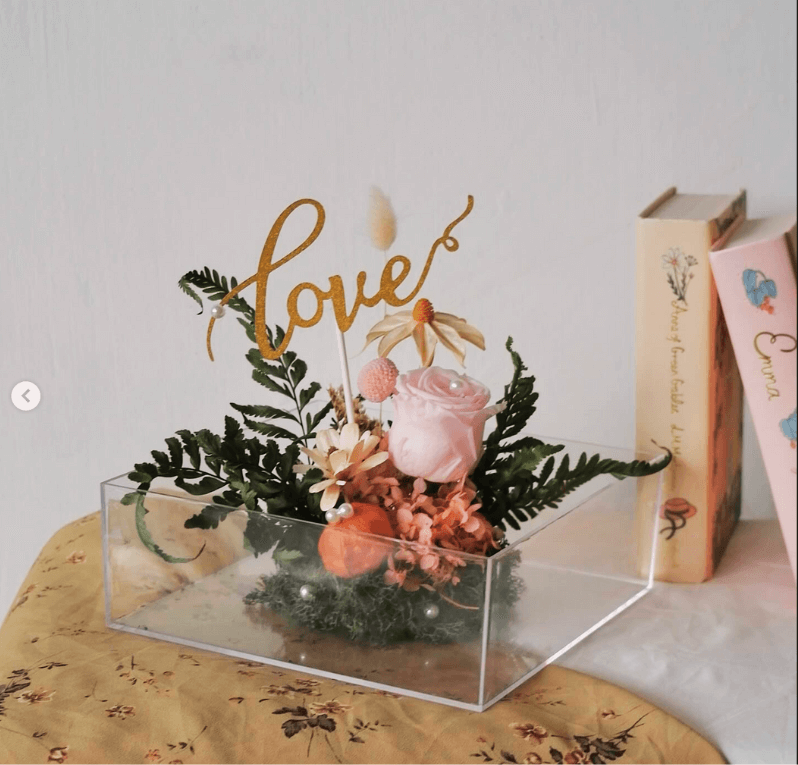 Preserved Valentine’s Day flower arrangement in a clear box, featuring a pink rose, dried blooms, and a golden “love” topper.
